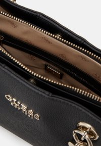 Musta Guess-käsilaukku kullanvärisillä vetoketjuilla, vaaleanruskea vuori, jossa on toistuva logokuosi, sekä kultainen brändilogo sisätaskussa.