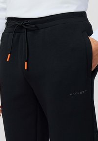 Zwarte sweatpants van zacht materiaal, met een elastische tailleband met een trekkoord en oranje tips, en een subtiel logo aan de zijkant.