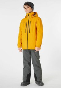 Giacca impermeabile gialla con cappuccio, cerniere nere e tasche; abbinata a pantaloni grigi con dettagli neri e stivali neri robusti.