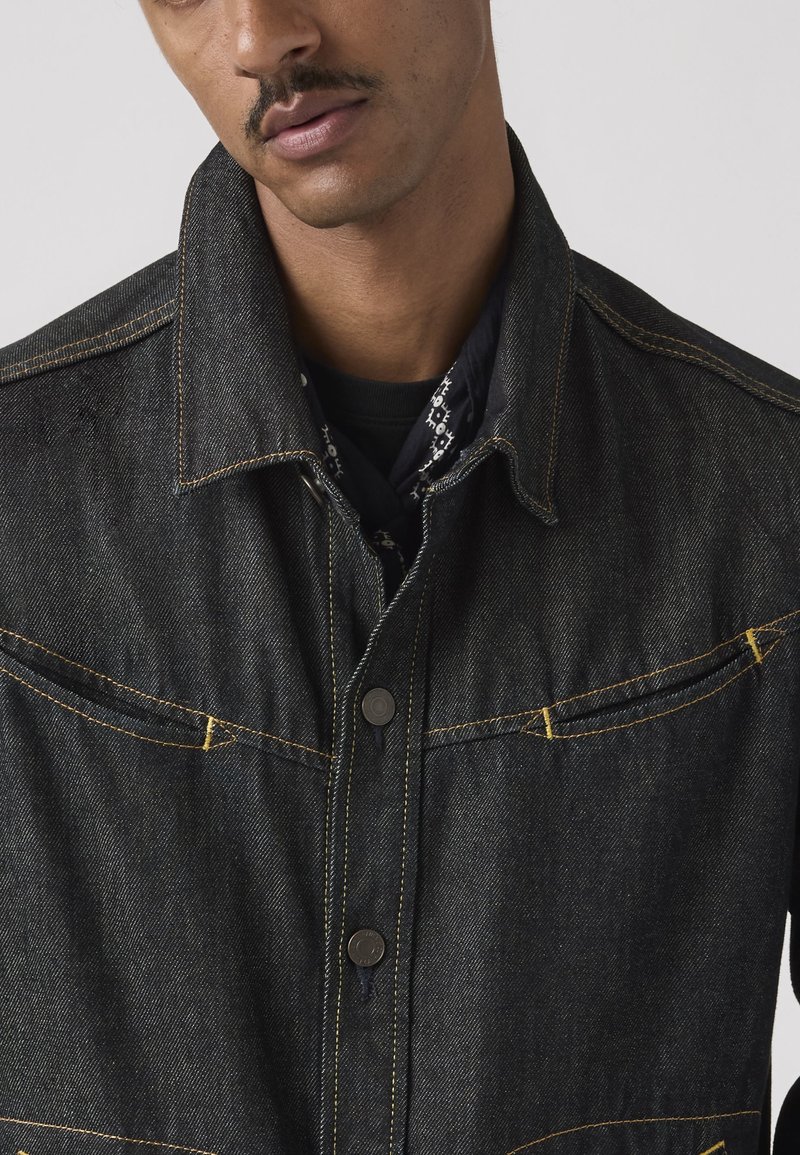 Mand iført en mørk denimjakke med gule syninger og et sort mønstret tørklæde om halsen mod en ensfarvet baggrund.