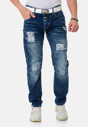 Blaue Jeans aus Denim mit ausgefransten Patches und fransigen Details, ausgestattet mit einem Knopfverschluss und abgetragenem Muster, kombiniert mit einem weißen Gürtel.