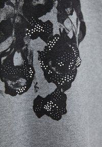 Harmaa kangas, jossa on musta kukkakuosi, korostettuna kiiltävillä strasseilla. Suunnittelu sisältää tekstuurin, joka on sekä sileä että kankaan luonteenomainen.