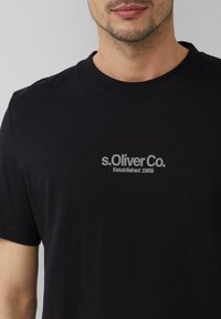 Czarny bawełniany t-shirt z okrągłym dekoltem. Z przodu znajduje się szary napis "s.Oliver Co. Established 1969". Miękka tekstura, krótkie rękawy.