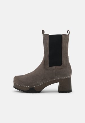 Bottine en daim gris à talon bloc, avec panneau latéral élastique noir, bout arrondi et languettes de tirage sur fond blanc.