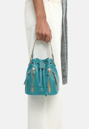 Ręka trzyma małą, turkusową, skórzaną torebkę typu bucket z wieloma przednimi kieszeniami na zamek, niebieskim ściągaczem i krótkim uchwytem na tle białych spodni.