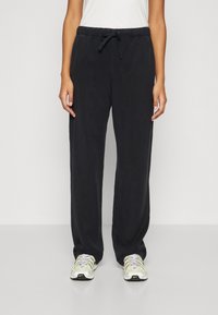 Black, relaxed-fit trousers with an elastic waistband, drawstring closure, and a smooth texture, paired with white and yellow sneakers.