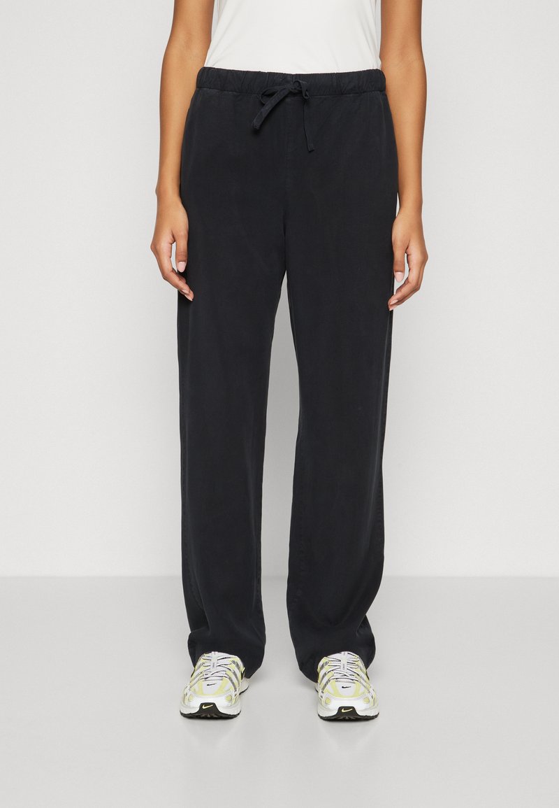 Black, relaxed-fit trousers with an elastic waistband, drawstring closure, and a smooth texture, paired with white and yellow sneakers.