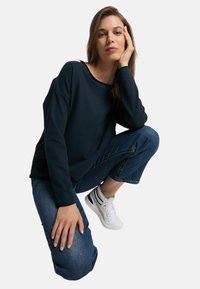 Navy long-sleeve sweatshirt with a loose fit, round neckline, and ribbed cuffs, paired with blue cropped jeans and white sneakers.