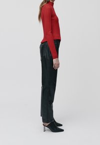 Femme de profil portant un col roulé à bretelles rouges, un pantalon en cuir noir et des mules à talons noirs sur fond blanc.