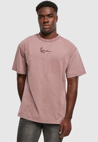 Heledast roosa puuvillane lühikeste varrukatega T-särk ümmarguse kaelusega ja õrn must tikand rinnal. Kangas näib olema veidi struktureeritud.