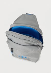 Γκρι υφασμάτινο σακίδιο με μπλε εσωτερικό, κλείσιμο με φερμουάρ, μπροστινή τσέπη και εμφανές λογότυπο σε μπλε κέντημα. Λεπτή υφή, συμπαγής σχεδίαση.