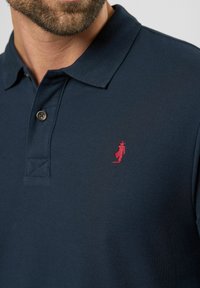 Marineblå polo shirt lavet af struktureret stof med et rødt broderet logo på brystet og to brune knapper ved kraven.