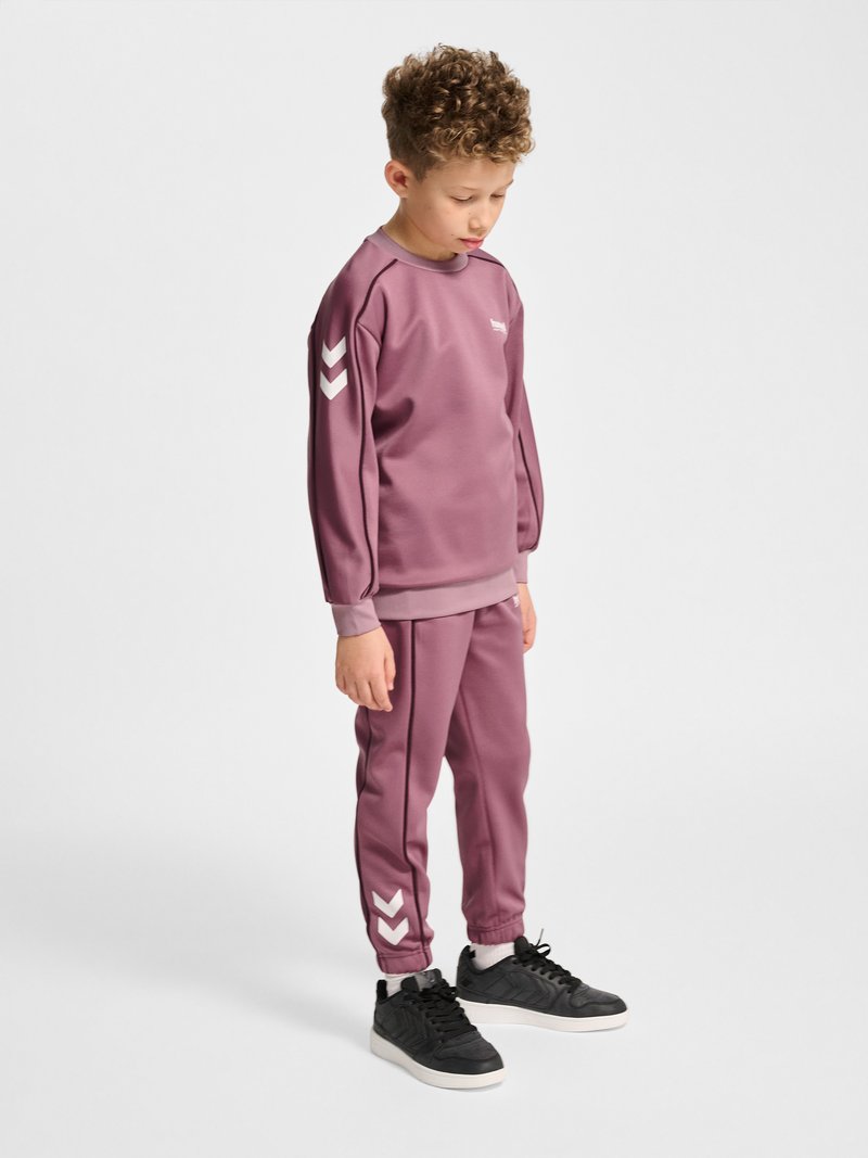 Dziecko z kręconymi włosami ubrane w mauve dres z białymi pasami w kształcie jodełki, patrzące w dół, stojące na jednolitym białym tle.