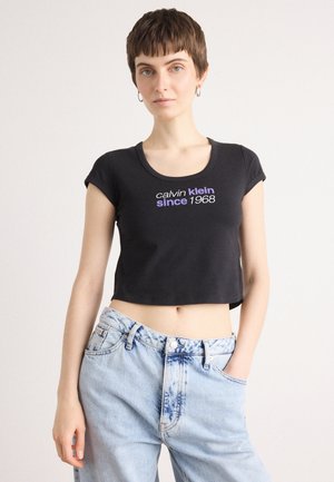 Zwarte cropped t-shirt met korte mouwen en ronde hals. De voorkant heeft witte en paarse tekst met de tekst "calvin klein since 1968." Gecombineerd met lichtblauwe jeans.
