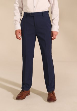 Uomo con pantaloni da abito blu navy, camicia bianca a maniche lunghe e scarpe eleganti in pelle marrone che sta in piedi su un pavimento beige.