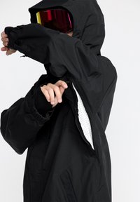 Veste noire imperméable avec capuche, dotée d'une fermeture éclair apparente, de poignets élastiques et d'une doublure en mesh. Le design met l'accent sur la fonctionnalité et le confort.