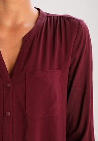 Primo piano di una persona che indossa una blusa bordeaux con bottoni, dotata di tasca sul petto e dettagli arricciati sulle spalle.