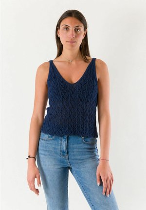 Donna con lunghi capelli castani che indossa un top smanicato in maglia blu navy e jeans blu, in piedi davanti a uno sfondo chiaro e semplice.