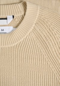 Pulover bej din tricot, cu o textură ribbed, guler rotund și o etichetă vizibilă la guler care indică informațiile despre brand și mărime.