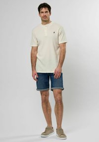 Kremfarget kortermet henley-skjorte med tre knapper, brukt med rullet denimshorts og beige sneakers. Enkel design og teksturert stoff.
