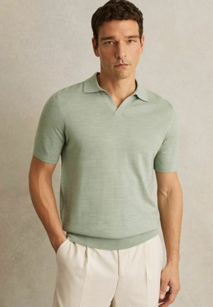 Cămașă polo verde deschis, cu mâneci scurte, având un decolteu în formă de V și tiv ribbat. Purtați cu pantaloni bej, evidențiind o textură moale.