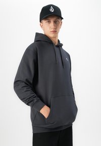 Young man wearing a dark gray hoodie with front pocket and black cap with white logo, standing against white background.