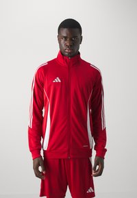Rød atletisk træningsdragt lavet af glat stof med hvide detaljer, der har en høj krave, lynlås og tre striber på ærmerne.