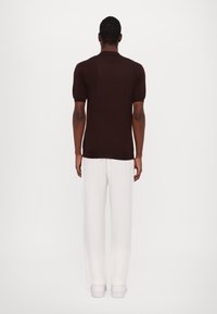 Hnědá polokošile s krátkým rukávem vyrobená z pletené tkaniny, v kombinaci s bílými širokými kalhotami a bílými teniskami, pohled zezadu.