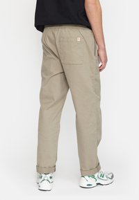 Beige broek met een elastische tailleband, twee achterzakken, opgerolde boorden en een klein label. Dragen met witte en groene sneakers.