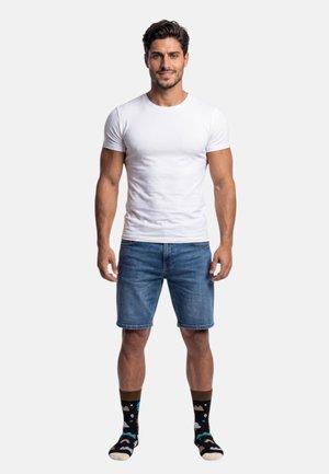 Mand med mørkt hår iført en tætsiddende hvid t-shirt, blå denimshorts og sorte sokker med farverige mønstre, stående mod en hvid baggrund.