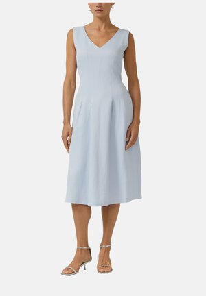 Femme portant une robe midi bleu clair sans manches à col en V, associée à des talons argentés avec bride à la cheville, debout devant un fond uni.