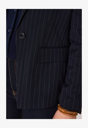 Marineblauer Nadelstreifen-Blazer mit Knopf und Pattentasche, getragen über einem dunklen Oberteil und Jeans, accessoriert mit braunen Wickelarmbändern am Handgelenk.