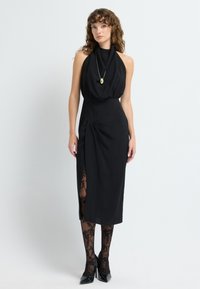 Czarna sukienka na halter z drapowanym dekoltem, dopasowanym w pasie i bocznym rozcięciem, które odsłania rajstopy z koronkowym wzorem. Dodatki obejmują złoty wisiorek.