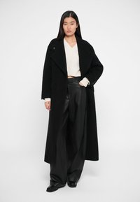 Melna vilnas mētelis ar platām atlokām un priekšējām kabatām, valkāts virs baltas adītas džempera un melniem mākslīgās ādas platiem biksēm.