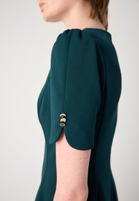 Teal dress featuring puff sleeves with scalloped edges and three gold button accents at the cuff. Smooth fabric with a fitted silhouette.