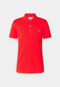 Červené polo tričko vyrobené z bavlny, s klasickým límečkem, zapínáním na dvě knoflíky, krátkými rukávy a zeleným logem Lacoste na hrudi.