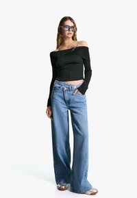 Black off-the-shoulder ribbed top paired with wide-leg light blue jeans. Model wears transparent sunglasses and silver footwear.