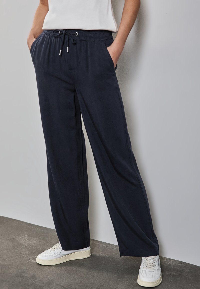 Personne portant un pantalon noir ample à cordon, des baskets blanches et une chemise blanche, debout contre un mur clair.