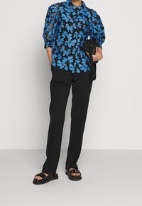 Black floral blouse with blue pattern, long sleeves, and a collar. Paired with black trousers and chunky black sandals, holding a round black bag.