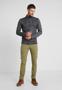 Giacca zip con motivo marmorizzato nero e grigio, con colletto alto, abbinata a pantaloni cargo verde oliva dotati di tasche laterali e stivali in cuoio marrone con lacci.