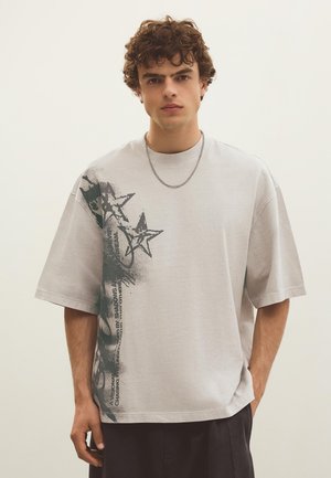 Mladić s kovrčavom kosom nosi svijetlosivu široku majicu s crnom zvjezdicom na lijevoj strani i crne hlače.