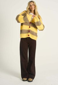 Žena s dlhými blond vlasmi, oblečená v žlto-hnedom pruhovanom huňatom kardigáne, tmavých širokých nohaviciach a béžových topánkach stojaca na jednofarebnom pozadí.