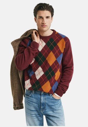 Rot- und cremfarbener Argyle-Pullover mit grünen, blauen und orangefarbenen Rauten. Weicher Stoff, gerippte Bündchen, getragen mit einer braunen Fischgrätjacke und blauen Jeans.