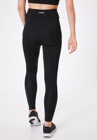 Zwarte hoge taille leggings met een gladde textuur, voorzien van reflecterende accenten langs het onderbeen, gecombineerd met zwarte sportschoenen.
