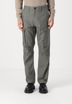 Homme portant un pantalon cargo gris avec des poches à rabat et des bottes noires, debout devant un fond clair uni.