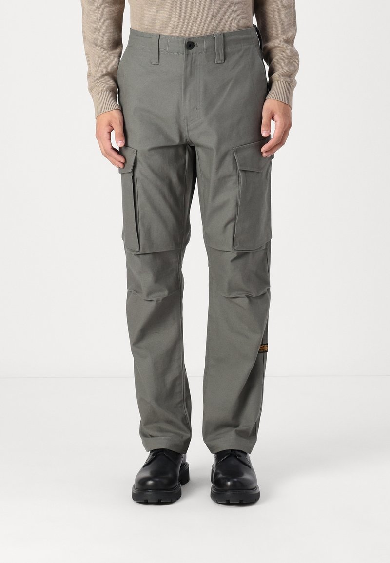 Homme portant un pantalon cargo gris avec des poches à rabat et des bottes noires, debout devant un fond clair uni.
