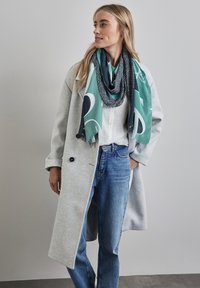 Grün und marineblau gemusterter Schal, der über einem weißen Hemd drapiert ist, kombiniert mit einem hellgrauen Mantel und blauen Jeans. Bemerkenswerte Texturen und Schichtung.