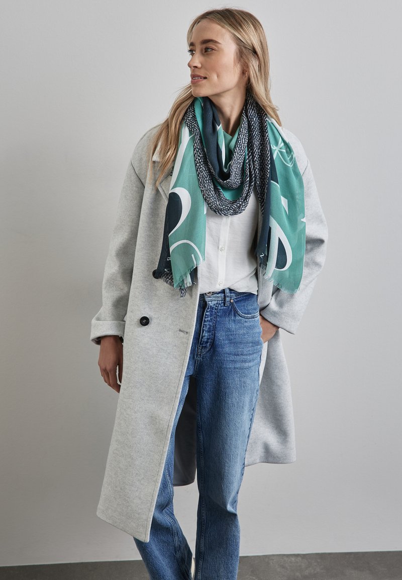 Grün und marineblau gemusterter Schal, der über einem weißen Hemd drapiert ist, kombiniert mit einem hellgrauen Mantel und blauen Jeans. Bemerkenswerte Texturen und Schichtung.