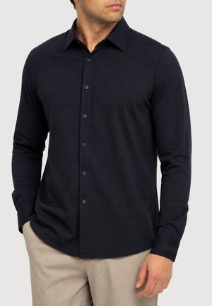 Uomo che indossa una camicia blu scuro tinta unita a maniche lunghe con bottoni e pantaloni beige, con una mano in tasca e guarda leggermente di lato.