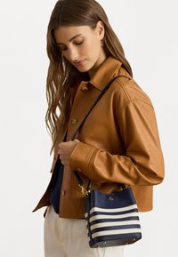 Saco balde azul-marinho com riscas creme, material texturizado, ferragens douradas e alça ajustável, combinado com uma jaqueta de couro castanho.